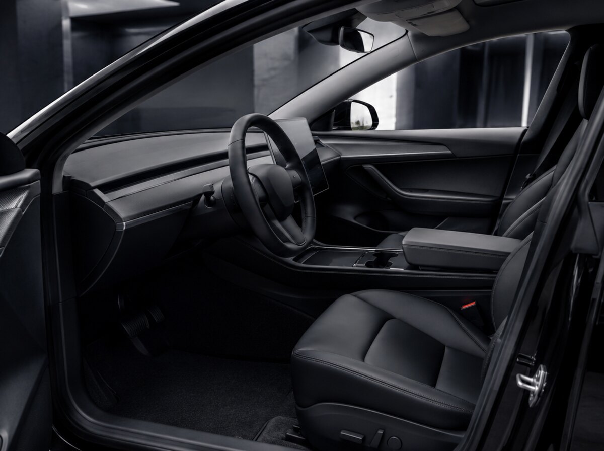 The front cabin of an electric sedan seen from the open driver door, black on black: black dash, black leather seats, black carpet, one landscape touchscreen mounted centrally with the screen dark, the car standing in a dim concrete structure.